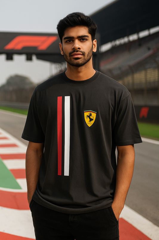 Indian  male model wearing a black Ferrari F1 t-shirt with vertical red and white racing stripes and Ferrari logo, standing confidently at a Formula 1 racetrack. Stylish, bold and modern apparel for F1 fans and streetwear lovers.