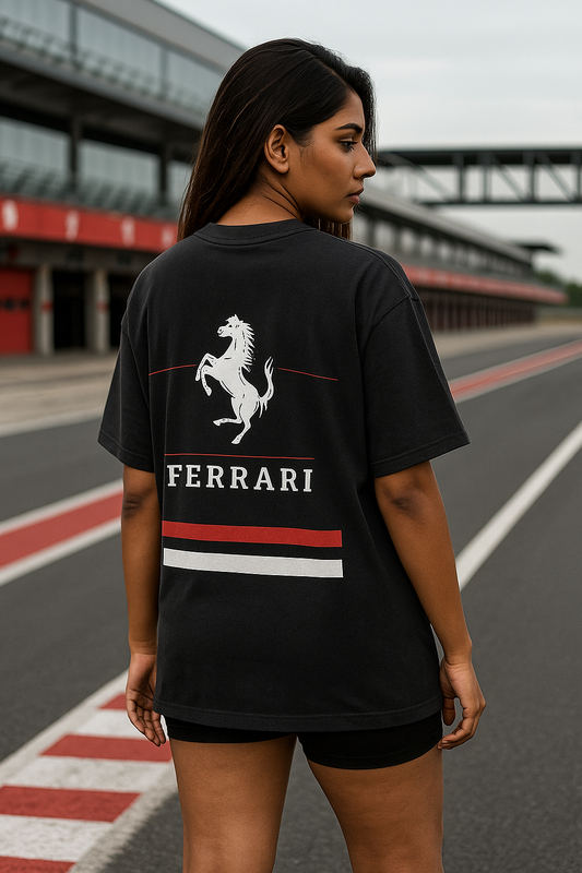 A confident Indian female model with fair skin and a fit build stands with his back to the camera, showcasing the back design of a black oversized Ferrari F1-themed t-shirt. The tee features a bold white prancing horse, red and white racing stripes, and the word "FERRARI" in capital letters. He is posed in a dramatic Formula 1 pit lane setting at sunset, with red lighting reflecting the Ferrari brand's energy and a blurred Ferrari sign in the background, creating a powerful and stylish racing vibe.