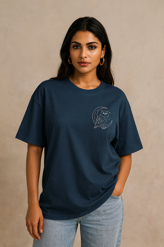 “Oversized unisex t-shirt featuring a majestic owl with glowing eyes perched on a crescent moon, surrounded by sacred geometry and stardust detailing on the front. The back showcases an owl in flight fading into mist, with mandala feather rings and the phrase ‘Wisdom Sees All’ above.