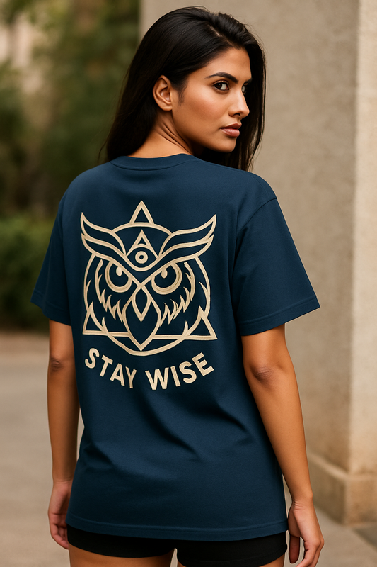 “Oversized unisex t-shirt featuring a majestic owl with glowing eyes perched on a crescent moon, surrounded by sacred geometry and stardust detailing on the front. The back showcases an owl in flight fading into mist, with mandala feather rings and the phrase ‘Wisdom Sees All’ above.
